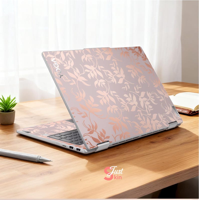 May include: A laptop with a floral design in rose gold and light pink. The word "YOGA" is visible on the top left corner of the laptop. The laptop is open on a wooden desk with a pen and small plant.