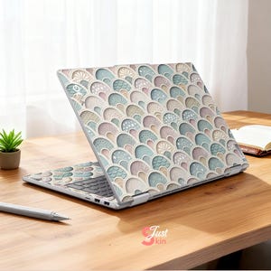 Puede incluir: Un ordenador port&aacute;til con una cubierta decorativa que presenta un patr&oacute;n repetitivo de formas de abanico en colores pastel. El dise&ntilde;o incluye tonos de azul, rosa y beige. El port&aacute;til est&aacute; abierto sobre una superficie de madera, con un bol&iacute;grafo y una peque&ntilde;a planta cerca.