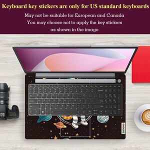 May include: Laptop with a space-themed keyboard skin showing planets, stars, and an astronaut. The keyboard has black keys and the screen displays a pink and white gradient. Text at the top indicates the stickers are for US keyboards.