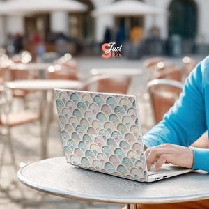 Puede incluir: Ordenador port&aacute;til con una cubierta decorativa con un patr&oacute;n repetido de formas de abanico en colores pastel. El port&aacute;til est&aacute; abierto sobre una mesa redonda, una persona escribe en el teclado. El logotipo de Just kin es visible.