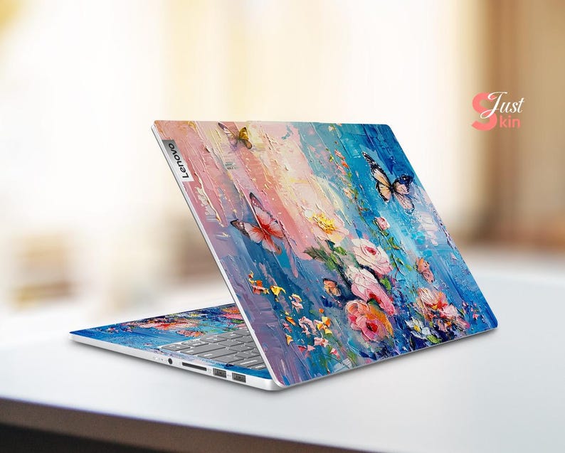 Puede incluir: Un ordenador port&aacute;til con un dise&ntilde;o floral y de mariposas colorido en la tapa. El dise&ntilde;o presenta colores rosa, azul, amarillo y verde. El port&aacute;til est&aacute; cerrado y el logotipo de Lenovo es visible en la tapa.