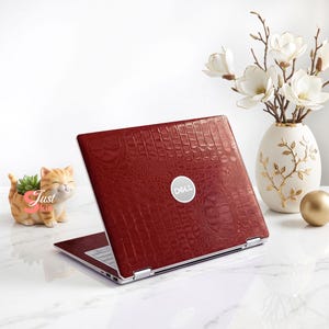 May include: A Dell laptop featuring a deep red, textured crocodile skin pattern on the outer lid. The laptop is positioned at an angle, displaying the silver Dell logo in the center of the lid. The surface of the laptop cover has a glossy finish.