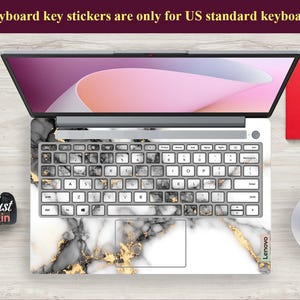May include: A white laptop with a black and gold marble design keyboard sticker. The sticker has the text "Lenovo" on the bottom right corner. The text "Keyboard key stickers are only for US standard keyboards" is at the top of the image.