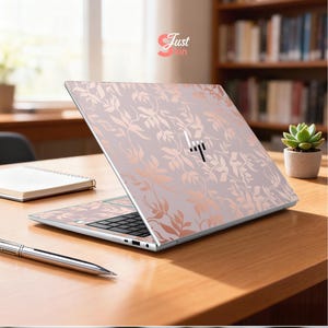 May include: A laptop with a floral design in shades of pink and rose gold. The laptop is open on a wooden desk, with a pen and notebook nearby. The HP logo is visible on the laptop.