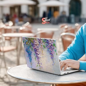 K&ouml;nnte beinhalten: Ein Laptop mit floralem Design mit lila, blauen und gr&uuml;nen Blauregenbl&uuml;ten. Der Laptop steht auf einem silbernen Tisch, und der Text "Just kin" ist sichtbar.