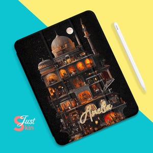 Puede incluir: Una funda negra para iPad con una ilustración detallada de un edificio de varios pisos con ventanas y luces. El edificio está sobre un fondo oscuro con una luna creciente y estrellas. El texto "Amelia" está escrito en letras doradas en la funda.