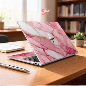 Op de afbeelding: Een laptop met een roze marmerontwerp en gouden accenten. De laptop is open en toont het toetsenbord en het scherm. Het merklogo is zichtbaar. De tekst "Just Skin" staat rechtsboven.