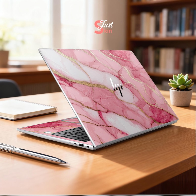 Op de afbeelding: Een laptop met een roze marmerontwerp en gouden accenten. De laptop is open en toont het toetsenbord en het scherm. Het merklogo is zichtbaar. De tekst "Just Skin" staat rechtsboven.