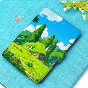 May include: A rectangular case featuring a vibrant illustration of a whimsical cottage nestled in a lush green landscape under a bright blue sky with fluffy white clouds. The case has a glossy finish and a black border.