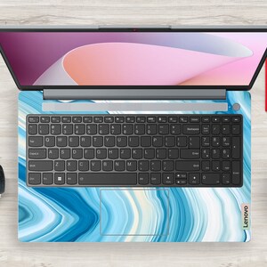 Op de afbeelding: Een laptop met een blauwe en witte marmer-design skin. De laptop heeft een zwart toetsenbord en een zilveren frame. Het scherm toont een roze en wit abstract ontwerp.
