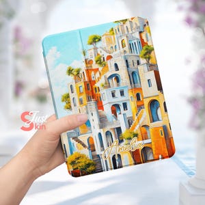 Puede incluir: Una funda para tablet colorida con una pintura de una ciudad con edificios apilados unos sobre otros. La funda tiene el nombre "Amelia" escrito en letras doradas.
