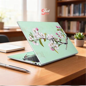 May include: A laptop with a light green cover adorned with a floral design of white and pink blossoms on a branch. The laptop is open on a wooden desk, with a pen and notebook nearby. The HP logo is visible.