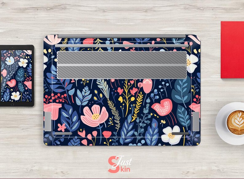 Op de afbeelding: Een laptop skin met een bloemenpatroon van roze, witte en gele bloemen en blauwe bladeren op een marineblauwe achtergrond. Het heeft een zilverkleurig ventilatiegedeelte. Een tablet met een bijpassend ontwerp is ook zichtbaar.