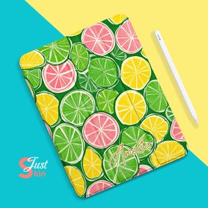 Pode incluir: Uma capa para iPad verde, amarela e rosa com um padrão desenhado à mão de limões e limas. A capa tem o nome "Amelia" escrito em letras douradas.