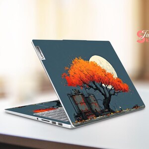 May include: A Lenovo laptop with a custom skin featuring an orange tree with a full moon in the background. The design includes industrial structures and a dark blue sky. The laptop is open, displaying the keyboard.
