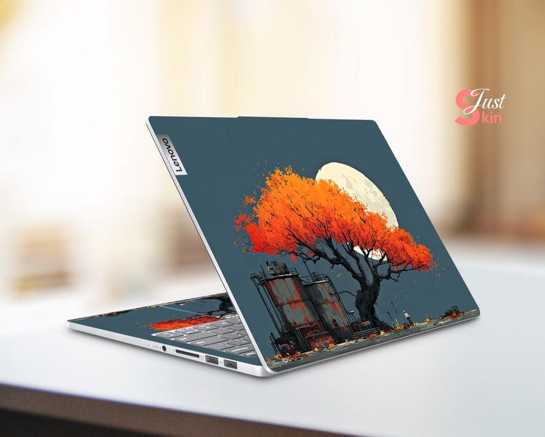 May include: A Lenovo laptop with a custom skin featuring an orange tree with a full moon in the background. The design includes industrial structures and a dark blue sky. The laptop is open, displaying the keyboard.