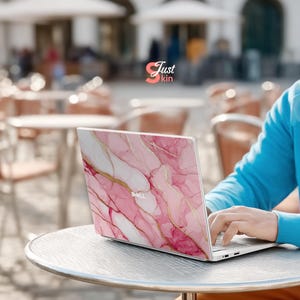 Op de afbeelding: Een Dell laptop met een roze en gouden marmeren ontwerp op de deksel. De laptop staat open op een metalen tafel. Een persoon in een blauwe trui typt op het toetsenbord. Het logo "Just kin" is zichtbaar op de achtergrond.