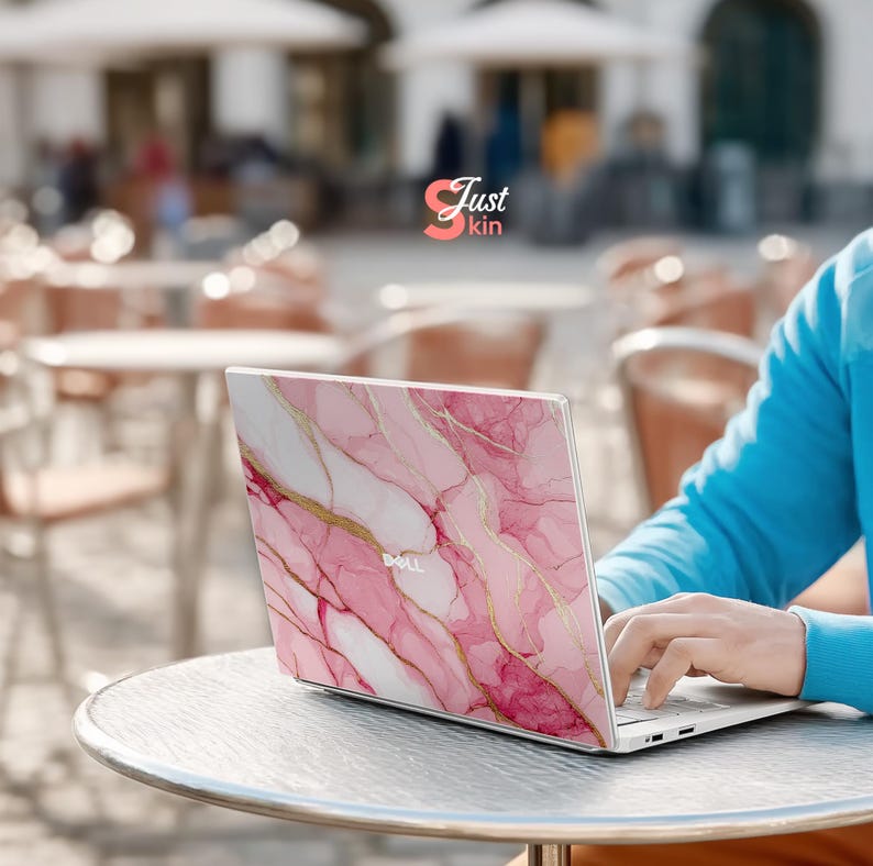 Op de afbeelding: Een Dell laptop met een roze en gouden marmeren ontwerp op de deksel. De laptop staat open op een metalen tafel. Een persoon in een blauwe trui typt op het toetsenbord. Het logo "Just kin" is zichtbaar op de achtergrond.