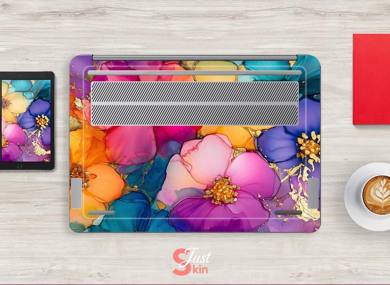 May include: A laptop skin featuring a vibrant floral design in shades of purple, pink, orange, and blue with gold accents. The design is abstract and artistic, with a watercolor effect. The laptop skin is on a wooden surface.