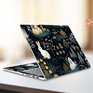 May include: A Lenovo laptop with a custom skin depicting a forest scene. The design features a white rabbit, plants, and a dark blue background with gold and white accents. The laptop is open, showing the keyboard.