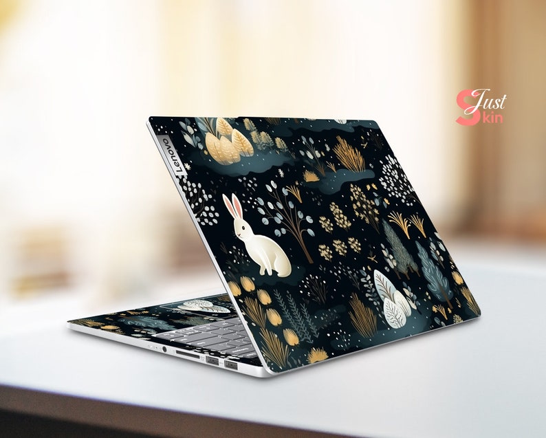 May include: A Lenovo laptop with a custom skin depicting a forest scene. The design features a white rabbit, plants, and a dark blue background with gold and white accents. The laptop is open, showing the keyboard.