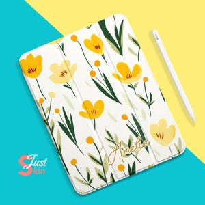 Könnte beinhalten: Weiße iPad-Hülle mit einem Blumenmuster mit gelben Blüten und grünen Blättern. Der Name "Amelia" ist in goldener Schrift auf der Hülle geschrieben.