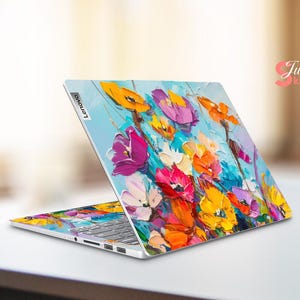 Puede incluir: Un ordenador port&aacute;til con un dise&ntilde;o floral colorido en la tapa. El dise&ntilde;o presenta una variedad de flores en tonos de rosa, morado, amarillo y naranja, pintadas en un estilo suelto y expresivo. El port&aacute;til est&aacute; cerrado y el logotipo de Lenovo es visible en la tapa.