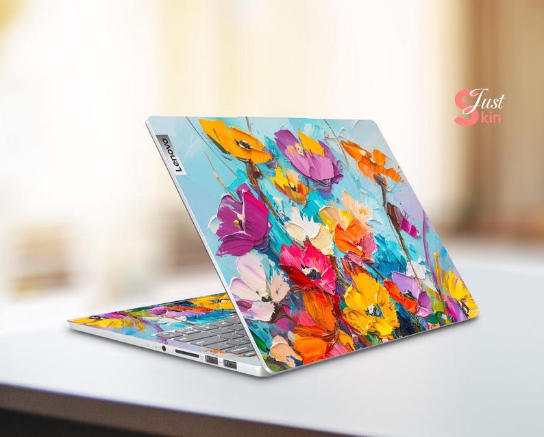 Puede incluir: Un ordenador port&aacute;til con un dise&ntilde;o floral colorido en la tapa. El dise&ntilde;o presenta una variedad de flores en tonos de rosa, morado, amarillo y naranja, pintadas en un estilo suelto y expresivo. El port&aacute;til est&aacute; cerrado y el logotipo de Lenovo es visible en la tapa.