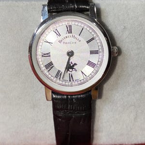 May include: A silver-toned wristwatch with a white face featuring black Roman numerals and the words "Beverly Hills Polo Club." The watch has a black leather band and a small image of a polo player. The watch is marked "Japan Movt."