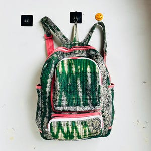 Könnte beinhalten: Ein gesteppter Rucksack mit grün-weißem Batikmuster, akzentuiert durch leuchtend rosa Paspeln und Reißverschlüsse. Der Rucksack verfügt über ein Hauptfach, eine kleinere Vordertasche und zwei Seitentaschen aus weichem, strukturiertem Stoff.