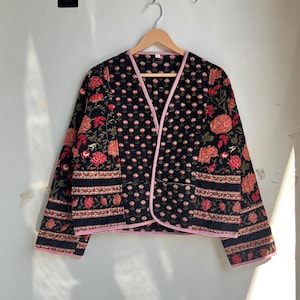 Op de afbeelding: Een zwarte jas met een bloemenpatroon in rood, oranje en groen. De jas heeft een roze rand langs de randen en een V-hals ontwerp. De jas hangt aan een houten hanger.