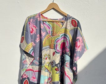 May include: A colorful, patterned caftan dress with a V-neck, hanging on a wooden hanger. The dress features a mix of floral and abstract designs in shades of gray, pink, yellow, and blue. The fabric appears lightweight and flowing, suitable for warm weather.