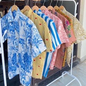 May include: A collection of button-down shirts in various patterns and colors hanging on a white rack. Designs include blue and white botanical prints, yellow stripes with red hearts, blue and white stripes, and floral patterns.