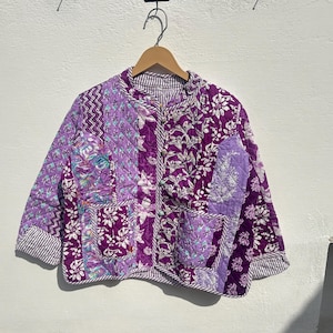 May include: A quilted jacket in shades of purple, featuring a patchwork design with floral and geometric patterns. The jacket has a button-up front, pockets, and striped trim. It hangs from a wooden hanger.