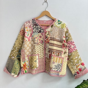 Puede incluir: Una chaqueta acolchada de patchwork con cierre de botón. La chaqueta está hecha de varias telas en tonos crema, rosa, verde y marrón. La chaqueta tiene un estampado floral y paisley.