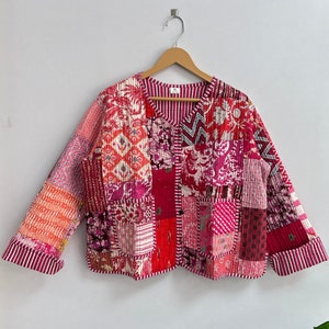 Puede incluir: Una chaqueta acolchada de patchwork rosa y roja con cuello en V y mangas largas. La chaqueta está hecha de una variedad de telas y tiene un aspecto único y hecho a mano.
