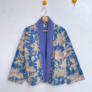 Può includere: Giacca con stampa floreale blu e beige con colletto e finiture blu a contrasto. La giacca è appesa a una gruccia di legno contro un muro bianco. La giacca ha un design ampio e aperto.