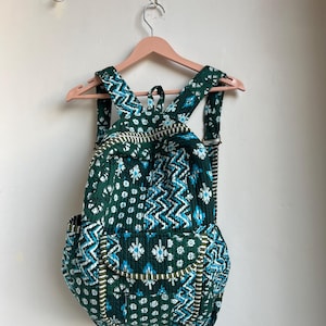 Può includere: Uno zaino trapuntato verde con un motivo floreale e geometrico bianco e blu. Lo zaino ha spallacci regolabili, una maniglia superiore e due tasche laterali. La borsa è appesa a un appendiabiti in legno rosa.