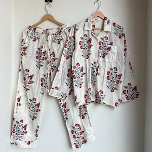May include: A two-piece pajama set with a floral pattern. The set includes a button-down shirt with a chest pocket and drawstring pants. The pajamas are white with a repeating pattern of red flowers and blue leaves.