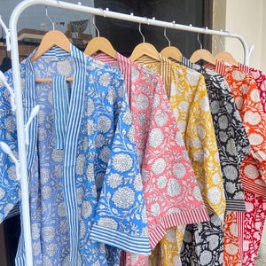 Puede incluir: Una colección de batas con estampados florales en varios colores, incluyendo azul, rosa, amarillo, negro y rojo, colgadas en un estante blanco. Cada bata presenta un diseño floral y un ribete diferentes.