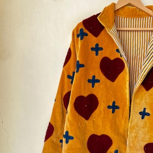 May include: Mustard yellow velvet jacket with a heart and cross pattern. The jacket features a red heart and blue cross design. The interior lining has a striped pattern. The jacket is hanging on a wooden hanger.