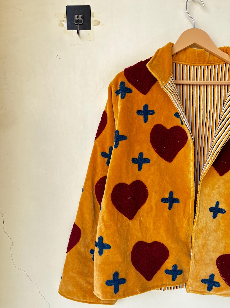 May include: Mustard yellow velvet jacket with a heart and cross pattern. The jacket features a red heart and blue cross design. The interior lining has a striped pattern. The jacket is hanging on a wooden hanger.