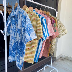 May include: A white metal clothing rack displays a variety of short-sleeved button-down shirts. The shirts feature diverse patterns, including blue and white tropical prints, yellow stripes with red hearts, and blue and white stripes. Other shirts have floral and geometric designs.