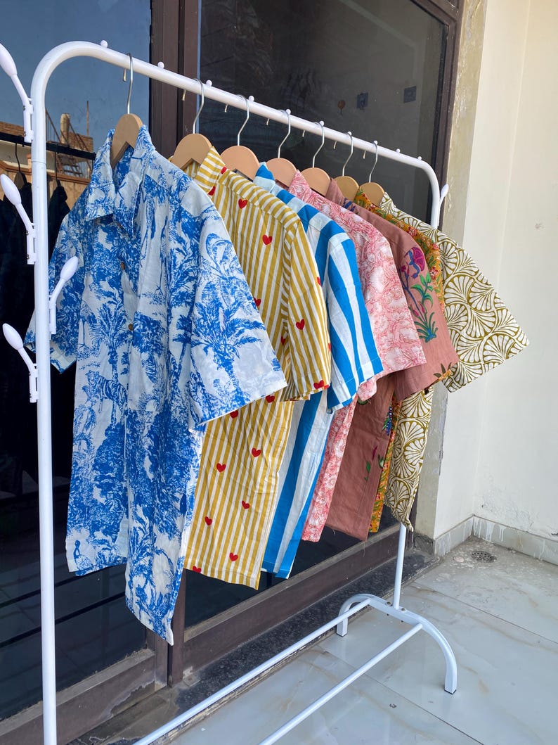 May include: A white metal clothing rack displays a variety of short-sleeved button-down shirts. The shirts feature diverse patterns, including blue and white tropical prints, yellow stripes with red hearts, and blue and white stripes. Other shirts have floral and geometric designs.