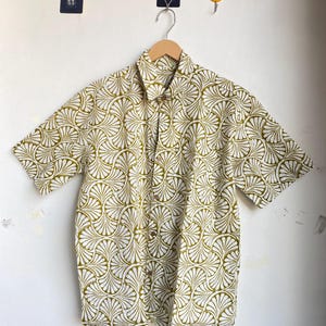 May include: A short-sleeved button-down shirt with a white background and a repeating olive green fan-shaped pattern. The shirt is hanging on a wooden hanger against a white wall.