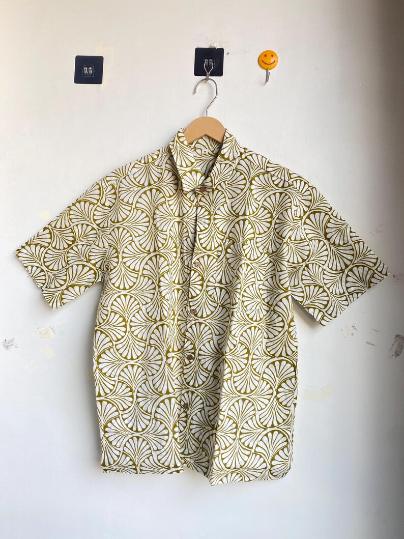 May include: A short-sleeved button-down shirt with a white background and a repeating olive green fan-shaped pattern. The shirt is hanging on a wooden hanger against a white wall.