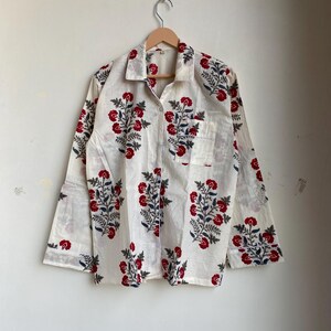 May include: White button-down shirt with a red and blue floral pattern. The long-sleeved shirt has a single pocket and is hanging on a wooden hanger. The floral design is repeated across the shirt.