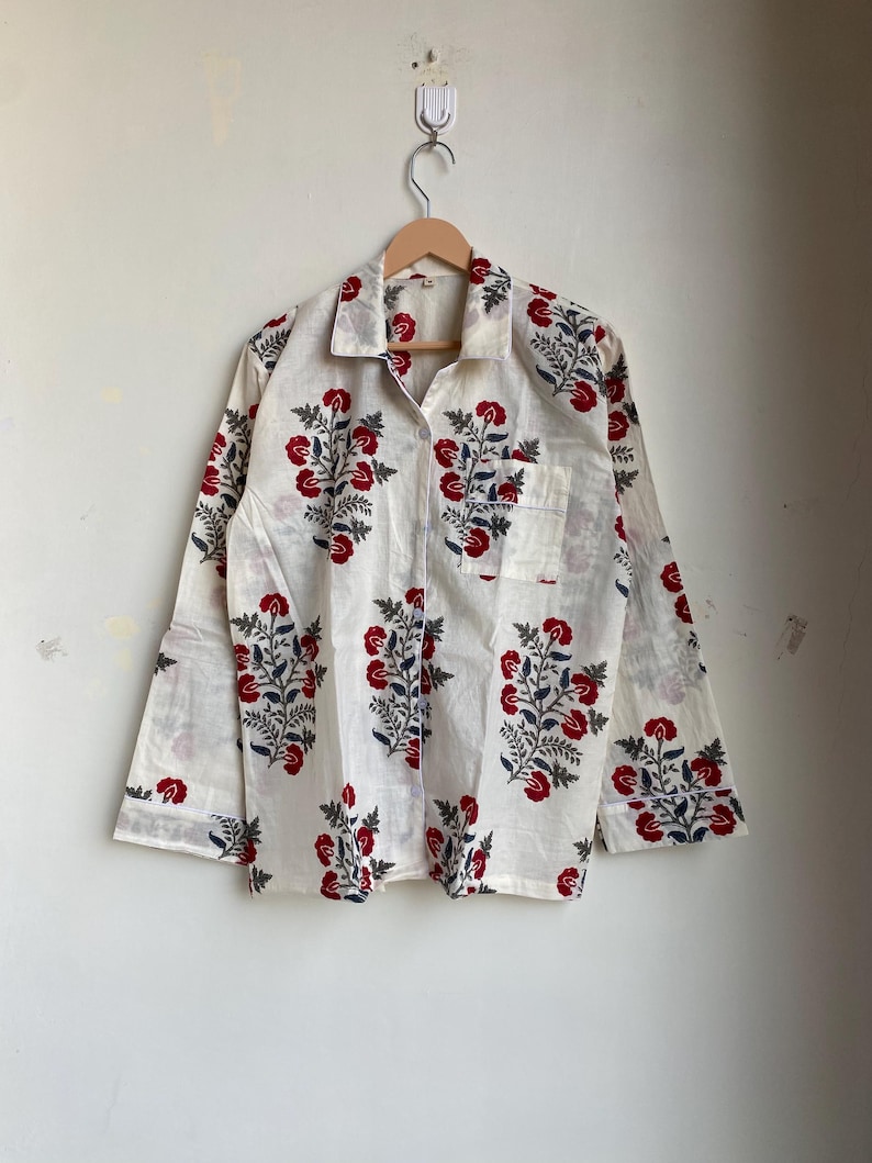 May include: White button-down shirt with a red and blue floral pattern. The long-sleeved shirt has a single pocket and is hanging on a wooden hanger. The floral design is repeated across the shirt.