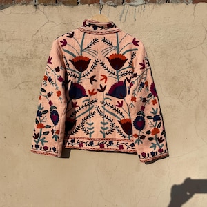 May include: A pink embroidered jacket with a floral pattern in shades of red, blue, and green. The jacket has a stand-up collar and long sleeves.