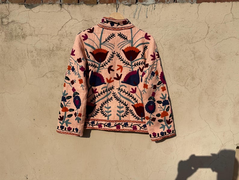 May include: A pink embroidered jacket with a floral pattern in shades of red, blue, and green. The jacket has a stand-up collar and long sleeves.